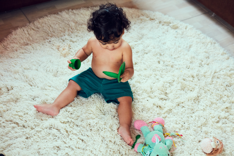 Uma criança está sentada em um tapete, brincando com uma xícara e uma tampa de plástico, ambos verdes. A imagem ilustra a matéria "Posicionamento da Fundação Maria Cecilia Souto Vidigal sobre o PL 1.338/2022, que propõe a regulamentação do ensino domiciliar".