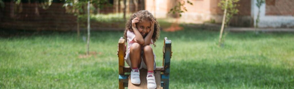 Menina sentada em brinquedo de madeira em um gramado, sorrindo durante uma brincadeira ao ar livre. Post internet segura.