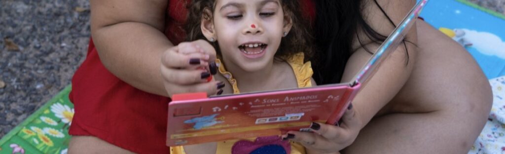 uma menina sorri enquanto lê um livro com ilustrações, acompanhada da mãe. a imagem ilustra o ganho da certificação CEBAS pela fundação maria cecilia souto vidigal.