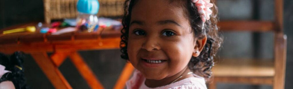 Retrato de uma menina negra sorridente usando uma tiara de flores rosa, em um ambiente de brincadeiras.